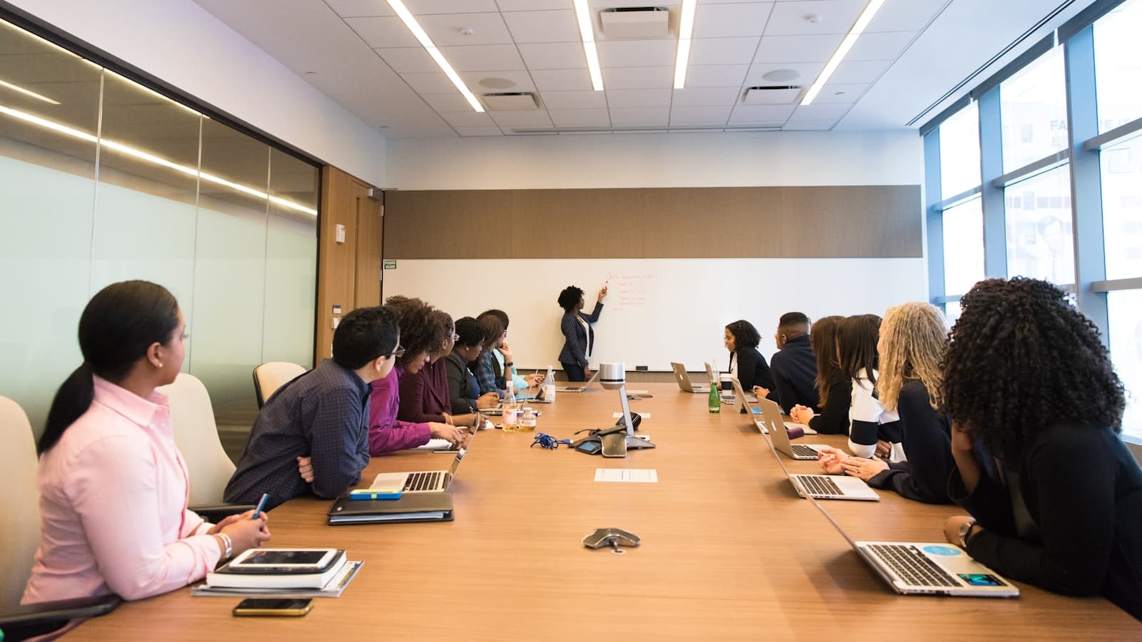 A group seated around a long conference table watching a colleague write on a whiteboard