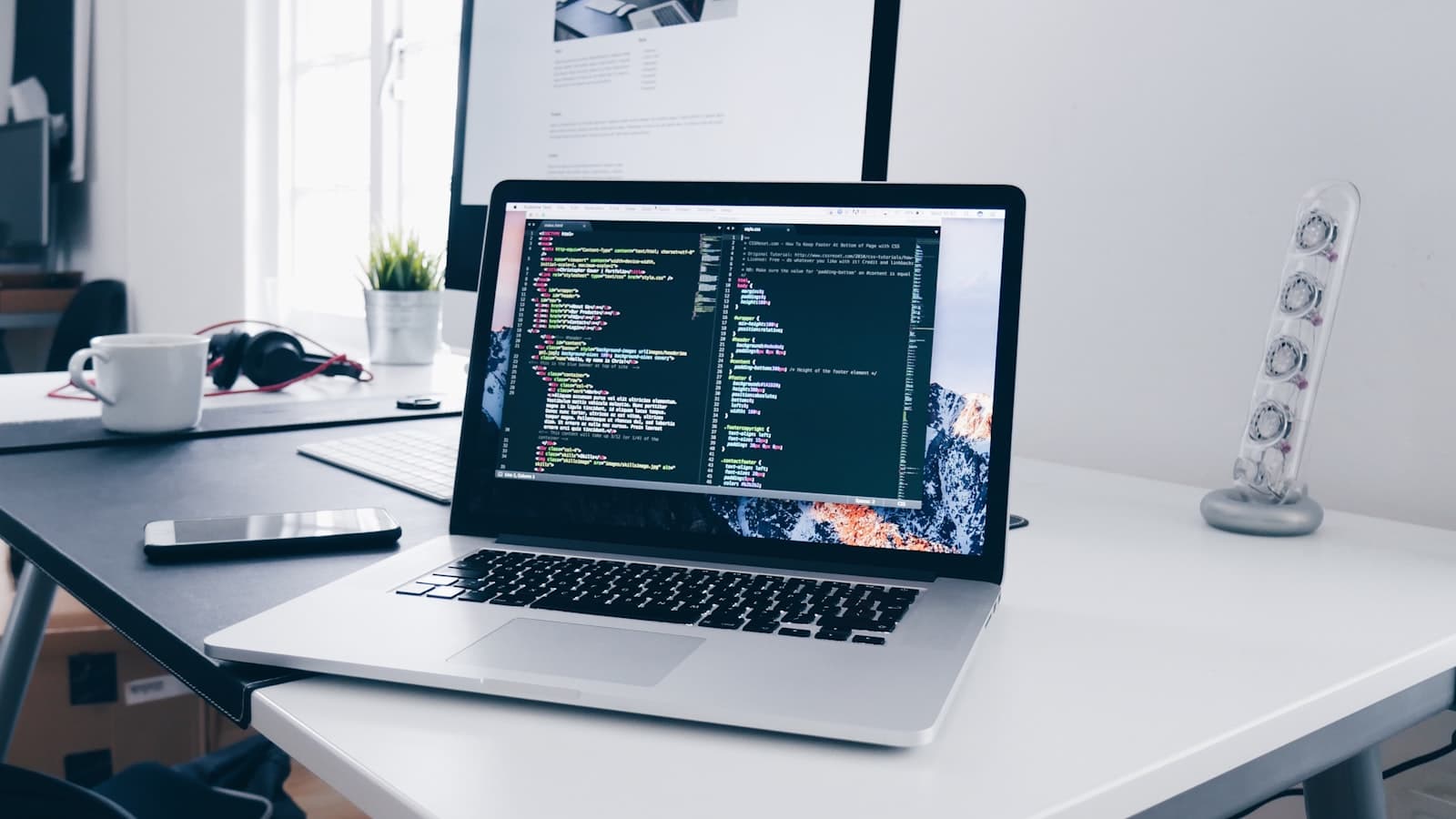A laptop showing code on a white desk beside a monitor, coffee cup and phone
