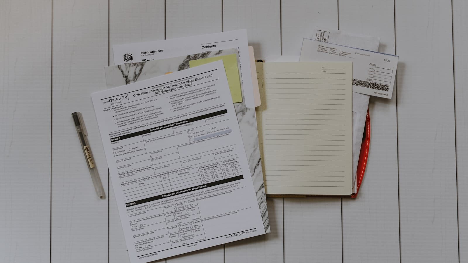 Printed tax forms, an open notebook and a pen laid out on a white surface