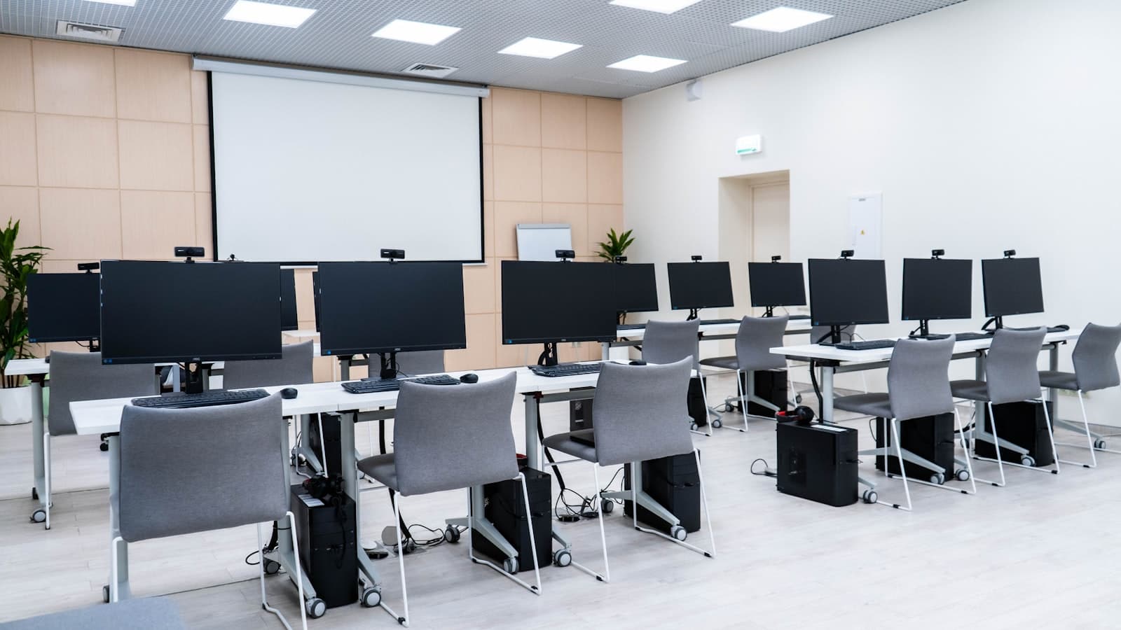 A classroom of desks with rows of desktop monitors, chairs and a projector screen