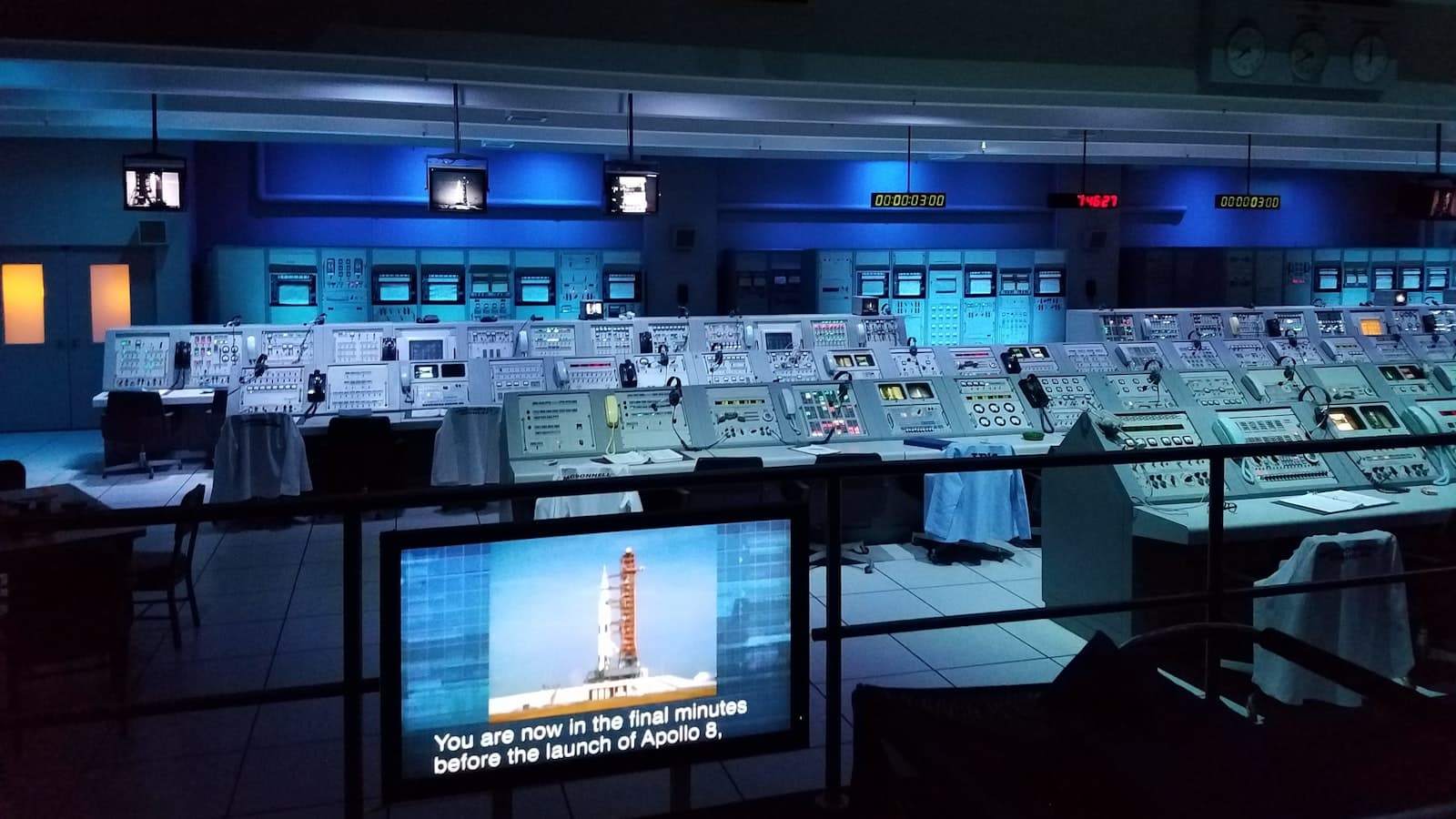 A dark historic mission control room with rows of consoles and a rocket launch on screen