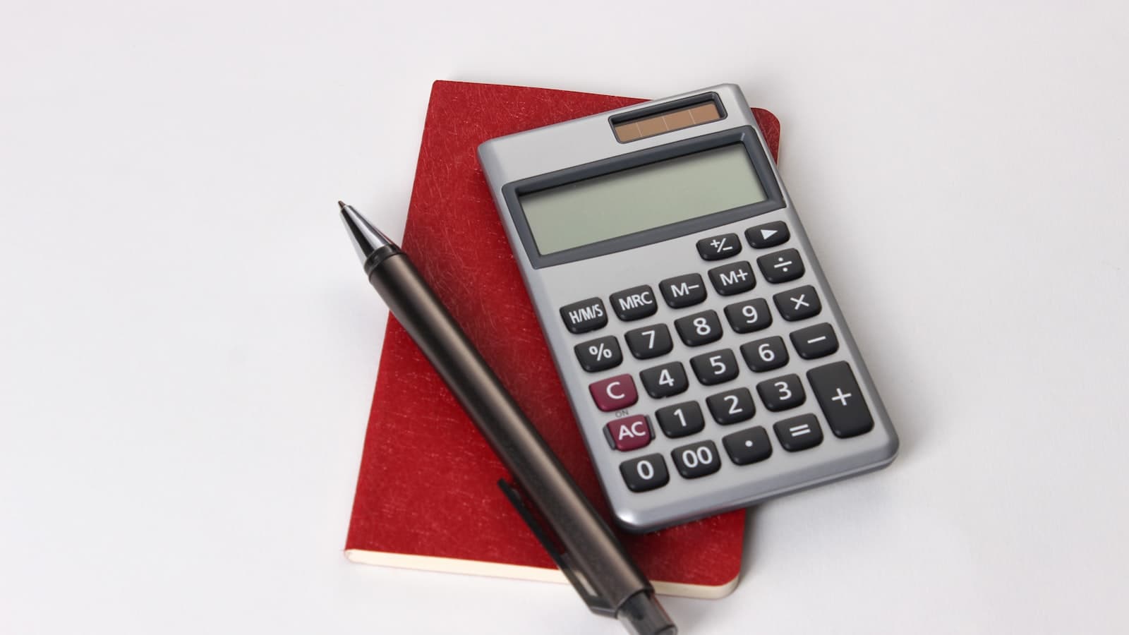 A grey calculator and a pen resting on a red notebook on a white surface